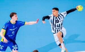 Ein Handballspieler in einem weiß-schwarzen Trikot bereitet sich auf den Wurf vor, während ein gegnerischer Spieler in einem blauen Trikot den Ball abwehrt, beide in Aktion auf einem blauen Spielfeld.