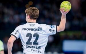 Ein Handballspieler, der ein weißes Trikot mit der Nummer 22 und dem Namen Andmann trägt, bereitet sich während eines Spiels auf den Wurf eines grünen Handballs vor. Der Spieler ist von hinten in einer Halle zu sehen.