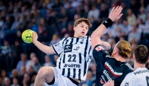 Ein Handballspieler in einem schwarz-weiß gestreiften Trikot springt, um den Ball zu werfen, während er von einem Gegner in einem schwarzen Trikot während eines Spiels verteidigt wird, während die Zuschauer im Hintergrund zusehen.