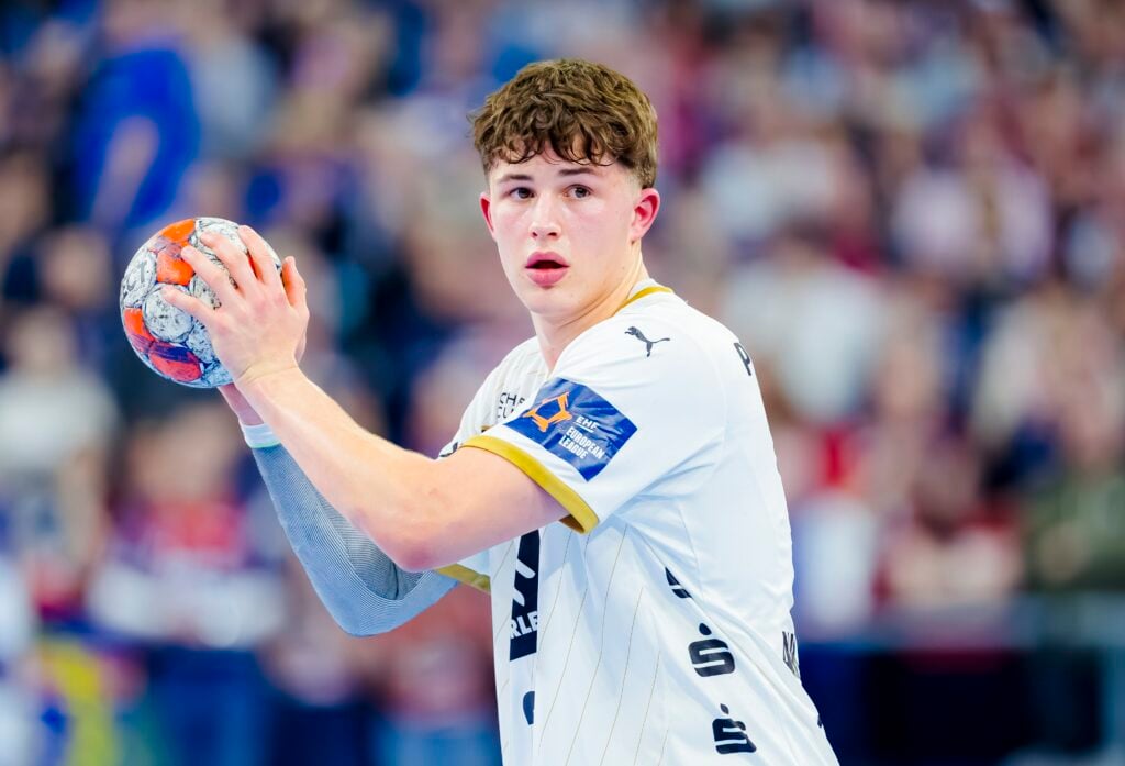 HANDBALL 2025/26: SG Flensburg-Handewitt - THW Kiel Ein junger männlicher Handballspieler in einem weißen Trikot hält einen Handball in der Hand und bereitet sich auf den Wurf vor, mit einem konzentrierten Gesichtsausdruck. Im Hintergrund ist eine unscharfe Menschenmenge zu sehen.