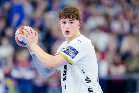 Ein junger männlicher Handballspieler in einem weißen Trikot hält einen Handball in der Hand und bereitet sich auf den Wurf vor, mit einem konzentrierten Gesichtsausdruck. Im Hintergrund ist eine unscharfe Menschenmenge zu sehen.