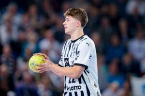Ein junger männlicher Handballspieler in einem schwarz-weiß gestreiften Trikot, der einen gelb-grünen Ball hält, steht auf einem Hallenplatz mit einer unscharfen Menschenmenge im Hintergrund.