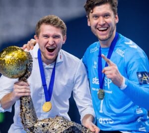 Zwei Männer, die mit Goldmedaillen und einem großen, kunstvollen Pokal feiern. Der eine trägt ein weißes Hemd, der andere ein blaues Sporttrikot, beide lächeln begeistert in die Kamera.