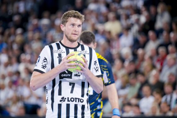 Ein Handballspieler in einem schwarz-weiß gestreiften Trikot hält den Ball und bereitet sich auf einen Spielzug während eines Spiels vor. Ein Spieler in einem blau-gelben Trikot steht hinter ihm, mit einer unscharfen Menge im Hintergrund.