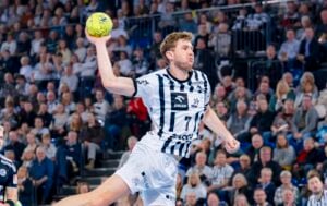 Ein männlicher Handballspieler in einem weiß-schwarz gestreiften Trikot springt in der Luft und bereitet sich darauf vor, einen gelben Ball zu werfen. Eine große Menschenmenge verfolgt das Spiel von den Tribünen im Hintergrund.