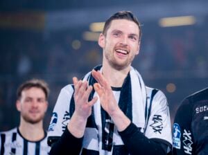 Ein männlicher Sportler in einem schwarz-weiß gestreiften Trikot lächelt und klatscht in die Hände, während er vor einer verschwommenen Menge steht. Ein anderer Spieler steht etwas hinter ihm, ebenfalls in Uniform.