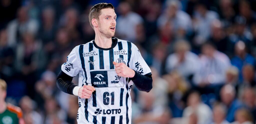 Ein männlicher Handballspieler in einem schwarz-weiß gestreiften Trikot mit der Nummer 61 steht auf dem Spielfeld, umgeben von einer unscharfen Menschenmenge im Hintergrund.