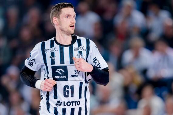 Ein männlicher Handballspieler in einem schwarz-weiß gestreiften Trikot mit der Nummer 61 steht auf dem Spielfeld, umgeben von einer unscharfen Menschenmenge im Hintergrund.