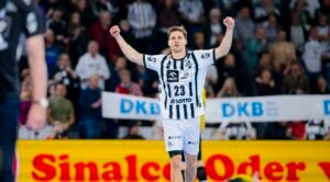 Ein männlicher Handballspieler in einem weiß-schwarz gestreiften Trikot hebt jubelnd beide Arme auf dem Spielfeld, während im Hintergrund eine jubelnde Menge und Werbebanner zu sehen sind.