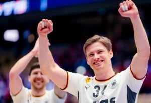 Ein lächelnder männlicher Sportler in einem weißen Trikot mit der Nummer 34 und der deutschen Flagge hebt jubelnd beide Arme. Ein anderer Spieler im Hintergrund hebt ebenfalls die Arme, beide zeigen Freude und Triumph.