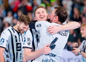 Zwei männliche Handballspieler in schwarz-weiß gestreiften Trikots umarmen sich und feiern fröhlich auf dem Spielfeld, während ein anderer Mitspieler im Hintergrund lächelt. Hinter ihnen sind eine jubelnde Menge und helle Lichter zu sehen.