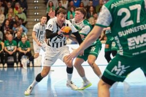 Ein Handballspieler in einem weiß-schwarz gestreiften Trikot stürmt mit dem Ball nach vorne und wird von einem Spieler in einem grünen Trikot eng verteidigt. Andere Spieler und Zuschauer sind im Hintergrund zu sehen.