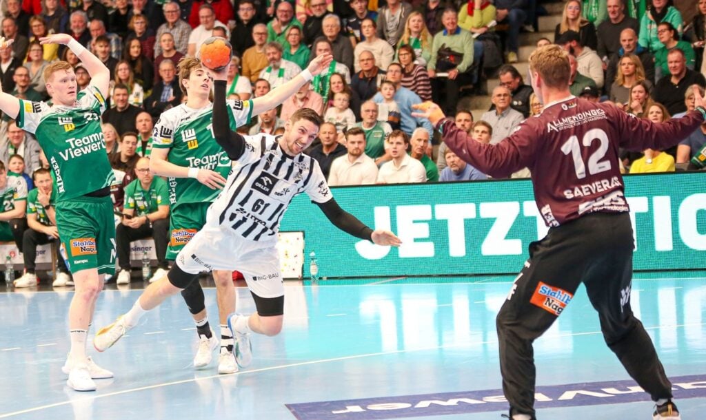 Ein Handballspieler in einem weiß-schwarzen Trikot springt, um den Ball in Richtung Tor zu schießen, während ein Torwart in einem kastanienbraunen Trikot sich darauf vorbereitet, den Ball zu blocken; zwei Verteidiger in Grün schauen von hinten zu. Die Tribünen sind voll mit Zuschauern.