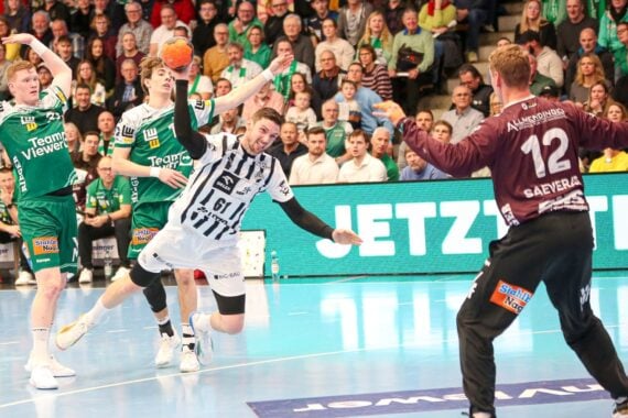 Ein Handballspieler in einem weiß-schwarzen Trikot springt, um den Ball in Richtung Tor zu schießen, während ein Torwart in einem kastanienbraunen Trikot sich darauf vorbereitet, den Ball zu blocken; zwei Verteidiger in Grün schauen von hinten zu. Die Tribünen sind voll mit Zuschauern.