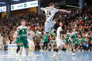 Ein Handballspieler in einem weiß-schwarzen Trikot springt in die Luft, um den Ball zu werfen, während seine Gegner in grün-gelben Trikots und eine dicht gedrängte Zuschauermenge das intensive Spiel in einer Halle verfolgen.