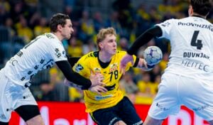 Ein Handballspieler in einem gelben Trikot versucht, den Ball zu passen, während er während eines intensiven Spiels von zwei Spielern in weißen Trikots eng verteidigt wird. Im Hintergrund ist eine unscharfe Menschenmenge zu sehen.