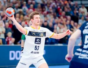 Ein männlicher Handballspieler in einem weißen Trikot mit der Nummer 53 bereitet sich darauf vor, den Ball während eines Spiels zu werfen. Der Hintergrund zeigt eine unscharfe Menschenmenge und einen anderen Spieler in einem dunklen Trikot.