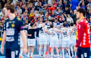 Eine Handballmannschaft in weißen Trikots versammelt sich und feiert in der Nähe des Tores, während Spieler in dunklen Trikots im Vordergrund stehen. Auf den Tribünen sitzen Zuschauer, die das Geschehen begeistert verfolgen.