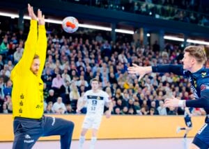 Ein Handballtorwart in einem gelben Trikot hebt die Arme, um einen Schuss abzublocken, während ein Spieler in einem blauen Trikot den Ball in Richtung Tor wirft. Eine große Menschenmenge verfolgt diesen intensiven Moment von der Tribüne aus.