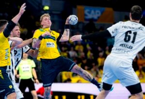 Ein tätowierter Handballspieler in einem gelben Trikot springt, um den Ball zu werfen, während gegnerische Spieler in weißen Trikots den Ball verteidigen.