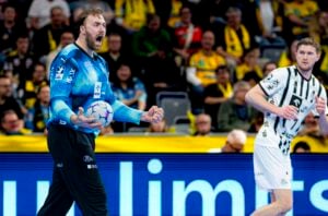 Ein Handballtorwart in einem blauen Trikot schreit energisch, während er den Ball hält, während ein anderer Spieler in einer weiß-schwarzen Uniform in der Nähe steht. Zuschauer in gelben Hemden schauen von der Tribüne im Hintergrund zu.