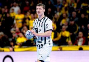 Ein Handballspieler in einem weiß-schwarz gestreiften Trikot hält einen blauen Ball und bereitet sich auf den Wurf vor, während im Hintergrund eine verschwommene Menschenmenge in gelben Hemden zu sehen ist.