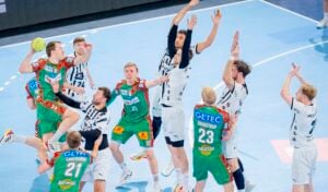 Eine Gruppe von männlichen Handballspielern in grün-weißen Trikots springt und greift während eines intensiven Spiels auf einem Hallenplatz nach dem Ball. Der Hintergrund zeigt einen blauen Boden und eine Teilansicht des Torraums.