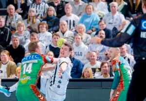 Ein Handballspieler in einem weißen Trikot versucht, ein Tor zu erzielen, während er von einem Spieler in einem grünen Trikot geblockt wird. Das Geschehen spielt sich vor den Augen zahlreicher Zuschauer ab, die von der Tribüne aus zusehen.