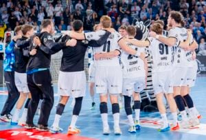 Eine Gruppe männlicher Handballspieler in weißen Trikots steht im Kreis auf einem blauen Spielfeld und legt die Arme umeinander. Ein kostümiertes Zebramaskottchen steht zwischen ihnen. Zuschauer füllen den Hintergrund.