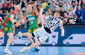 Ein Handballspieler in einem weiß-schwarzen Trikot springt, um den Ball zu werfen, während zwei gegnerische Spieler in grün-roten Trikots versuchen, den Ball zu verteidigen, während im Hintergrund eine Menge von Zuschauern zusieht.