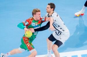 Zwei männliche Handballspieler kämpfen auf einem Hallenplatz um den Ball; der Spieler in Grün und Rot versucht vorzudringen, während der Spieler in Weiß eng verteidigt, wobei beide große Anstrengungen unternehmen.