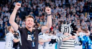 Ein lächelnder Mann mit feierlich erhobenen Armen steht vor einer Gruppe von Menschen und einem Zebra-Maskottchen, während im Hintergrund eine jubelnde Menge zu sehen ist.