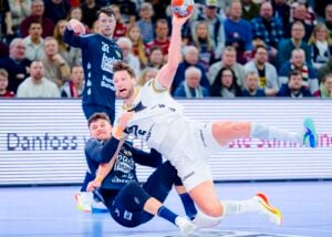 Ein Handballspieler in Weiß springt, um zu schießen, während er von einem Spieler in Blau angegriffen wird; ein intensiver Moment während eines Spiels mit einer konzentrierten Zuschauermenge im Hintergrund.
