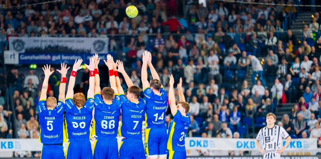 HANDBALL 2025/26: THW Kiel - VfL Gummersbach Eine Gruppe von Handballspielern in blauen Uniformen springt mit erhobenen Armen, um einen Schuss abzublocken, während ein Spieler in einer schwarz-weißen Uniform sich darauf vorbereitet, den Ball zu werfen. Der Hintergrund zeigt eine überfüllte Arena mit Zuschauern.