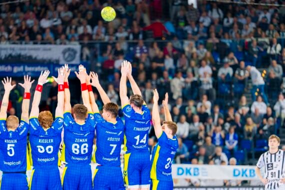 Eine Gruppe von Handballspielern in blauen Uniformen springt mit erhobenen Armen, um einen Schuss abzublocken, während ein Spieler in einer schwarz-weißen Uniform sich darauf vorbereitet, den Ball zu werfen. Der Hintergrund zeigt eine überfüllte Arena mit Zuschauern.