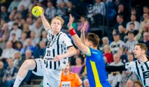 Ein männlicher Handballspieler in einem weiß-schwarzen Trikot springt, um den Ball zu werfen, während ein Verteidiger in einem blau-gelben Trikot die Arme zum Abwehren hebt. Ein Offizieller schaut genau zu, während die Menge zuschaut.