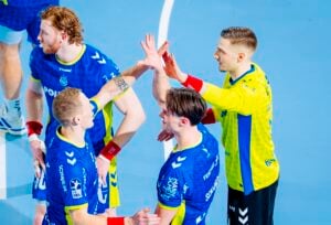 Vier männliche Handballspieler feiern auf einem Hallenplatz. Drei von ihnen tragen blaue Trikots, und einer in einem gelben Trikot gibt High-Fives. Sie sehen nach einem Spiel glücklich und engagiert aus. Der Hintergrund ist ein hellblauer Hallenboden.