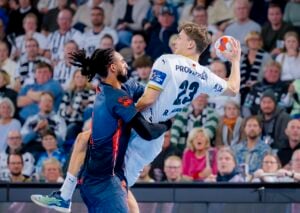 Zwei männliche Handballspieler liefern sich einen intensiven Wettkampf. Der eine in einem weißen Trikot (23) springt mit dem Ball, während der andere in einem dunkelblauen Trikot verteidigt. Eine Zuschauermenge beobachtet das Geschehen im Hintergrund.