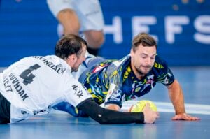 Zwei männliche Handballspieler tauchen und greifen nach einem gelben Ball auf einem Hallenplatz. Einer trägt ein weißes Trikot mit der Nummer 4, der andere ein blau gemustertes Trikot. Der Hintergrund ist unscharf.