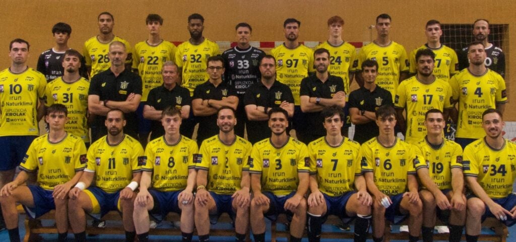 26_vor_Bidasoa_Team Eine Männer-Handballmannschaft stellt sich in einer Halle für ein Gruppenfoto auf. Die Spieler in gelben Trikots und blauen Hosen sitzen und stehen in Reihen, während Trainer und Betreuer in schwarzen Hemden in der Mitte stehen. Alle sind der Kamera zugewandt.