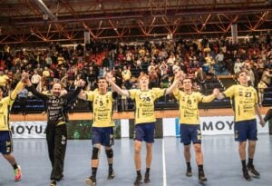 Eine Gruppe männlicher Handballspieler in gelben Trikots feiert, indem sie sich an den Händen halten und diese vor den jubelnden Fans in einer Sportarena gemeinsam hochheben.