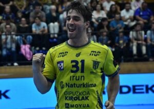 Ein männlicher Handballspieler in einem gelben Trikot mit der Nummer 31 jubelt auf dem Spielfeld und lächelt dabei enthusiastisch. Im unscharfen Hintergrund sind Zuschauer zu sehen.