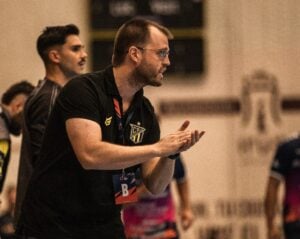 Ein Mann mit Brille und einem schwarzen Hemd mit einem CPR-Sportlogo steht in einem Raum, klatscht und scheint Anweisungen zu geben. Im unscharfen Hintergrund sind weitere Personen in Sportkleidung zu sehen.