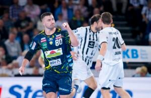 Ein männlicher Handballspieler in einem blau-gelben Trikot hebt jubelnd die Faust auf einem Spielfeld, während im Hintergrund zwei Gegner in weiß-schwarzen Trikots laufen. Auf der Tribüne sind Zuschauer zu sehen.