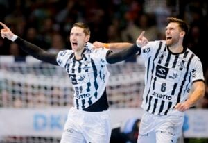 Zwei männliche Handballspieler in schwarz-weiß gestreiften Trikots feiern ausgelassen auf dem Spielfeld, wobei der eine nach vorne zeigt und der andere mit erhobenem Arm schreit. Im Hintergrund sind eine unscharfe Menge und ein Netz zu sehen.