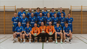 Eine Herren-Handballmannschaft sitzt in einer Halle auf Bänken und trägt blaue Trikots mit Nummern und e:zone-Logo; drei Personen in orangefarbenen Trikots halten grüne und weiße Bälle in der ersten Reihe. Hinter ihnen ist eine hölzerne Turnhallenwand zu sehen.