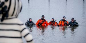 Fünf Taucher in Neoprenanzügen und orangefarbenen Trockenanzügen stehen hüfttief im Wasser und lächeln in die Kamera. Im Vordergrund ist teilweise eine verschwommene Gestalt in einem gestreiften Kostüm zu erkennen, die den Tauchern gegenübersteht.