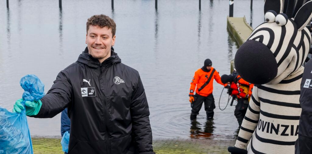 Handball: „För de Küste“-Projekt des THW Kiel Ein Mann in einer schwarzen Jacke mit einem blauen Müllsack steht neben einem Zebramaskottchen und zwei Personen in orangefarbenen Anzügen, die im flachen Wasser arbeiten. Sie scheinen an einer Umweltsäuberung beteiligt zu sein.