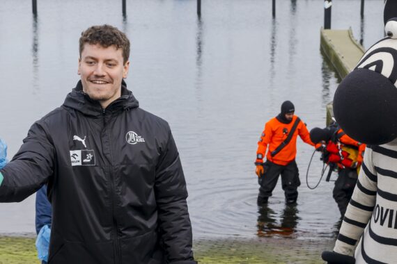 Ein Mann in einer schwarzen Jacke mit einem blauen Müllsack steht neben einem Zebramaskottchen und zwei Personen in orangefarbenen Anzügen, die im flachen Wasser arbeiten. Sie scheinen an einer Umweltsäuberung beteiligt zu sein.