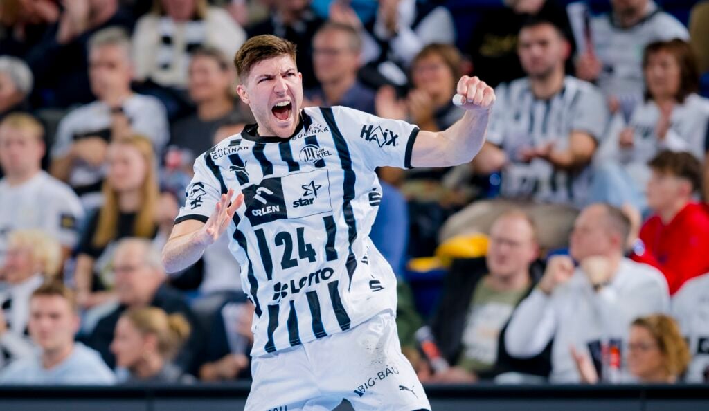 Ein männlicher Handballspieler in einem schwarz-weiß gestreiften Trikot jubelt energisch auf dem Spielfeld, während im Hintergrund eine begeisterte Menge steht.